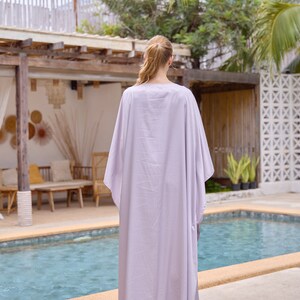 Puede incluir: Una mujer con un vestido largo y fluido de color lila claro tipo kaftan, de pie junto a una piscina.
