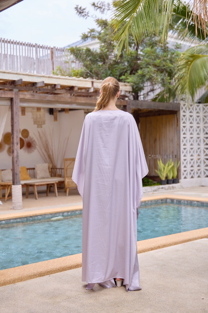 Puede incluir: Una mujer con un vestido largo y fluido de color lila claro tipo kaftan, de pie junto a una piscina.