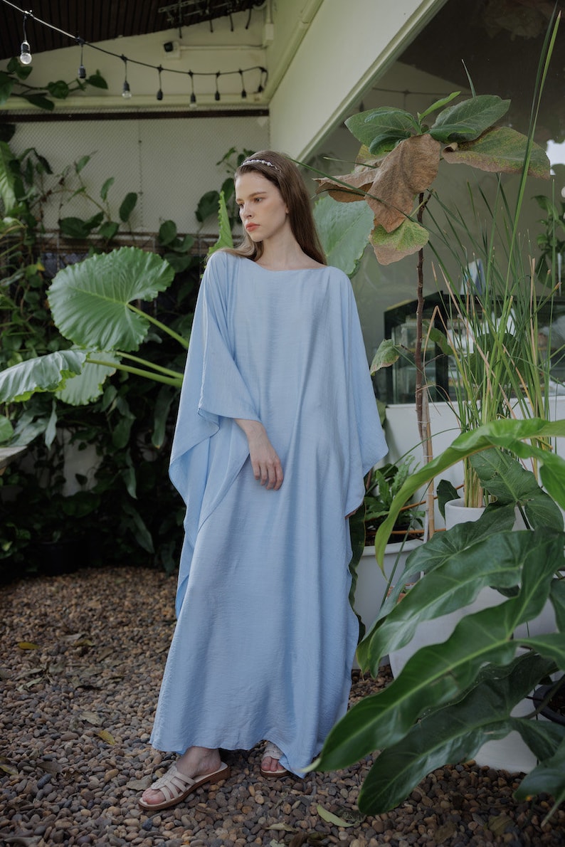 K&ouml;nnte beinhalten: Ein hellblaues Maxikleid mit einer lockeren, flie&szlig;enden Silhouette. Das Kleid hat einen runden Ausschnitt und lange &Auml;rmel. Das Model tr&auml;gt das Kleid mit Sandalen und einem Stirnband.