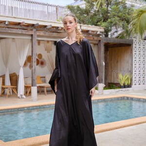Peut inclure: Longue robe caftan noire avec un col en V et des manches larges. L'ensemble comprend un pantalon noir assorti et des sandales blanches. L'arri&egrave;re-plan montre une piscine, un ciel nuageux et des palmiers.