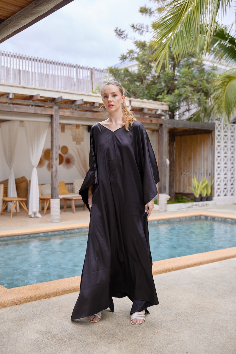 Peut inclure: Longue robe caftan noire avec un col en V et des manches larges. L'ensemble comprend un pantalon noir assorti et des sandales blanches. L'arri&egrave;re-plan montre une piscine, un ciel nuageux et des palmiers.