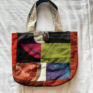 May include: A patchwork tote bag featuring a mix of colours and patterns, including red, orange, green, blue, and black. The bag has a single, brown button closure.