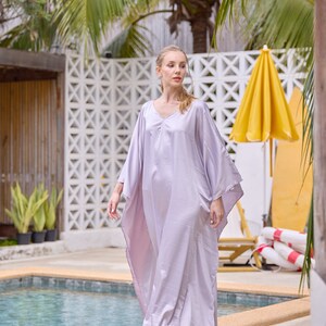 Puede incluir: Una mujer con un vestido kaftan de color lila claro y fluido junto a una piscina. El vestido tiene cuello en V y mangas largas.