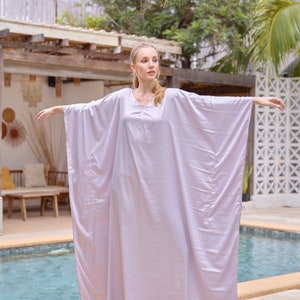 Puede incluir: Una mujer con un vestido largo y fluido de color lila claro estilo caft&aacute;n. Est&aacute; de pie junto a una piscina con los brazos extendidos.