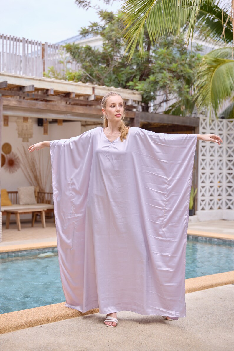 Puede incluir: Una mujer con un vestido largo y fluido de color lila claro estilo caft&aacute;n. Est&aacute; de pie junto a una piscina con los brazos extendidos.