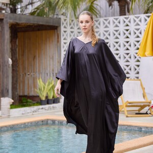 Peut inclure: Robe caftan noire, ample, avec un col en V et des manches larges. La personne porte des sandales blanches. En arri&egrave;re-plan, une piscine, des palmiers et un parasol jaune.
