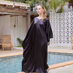Peut inclure: Longue robe caftan en satin gris fonc&eacute;, avec un col en V et des manches larges. La robe est longue et fluide, avec un &eacute;clat subtil. La personne porte des sandales blanches. L'arri&egrave;re-plan comprend une piscine et un feuillage tropical.
