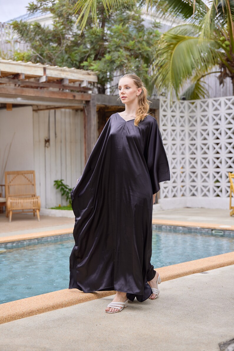 Peut inclure: Longue robe caftan en satin gris fonc&eacute;, avec un col en V et des manches larges. La robe est longue et fluide, avec un &eacute;clat subtil. La personne porte des sandales blanches. L'arri&egrave;re-plan comprend une piscine et un feuillage tropical.