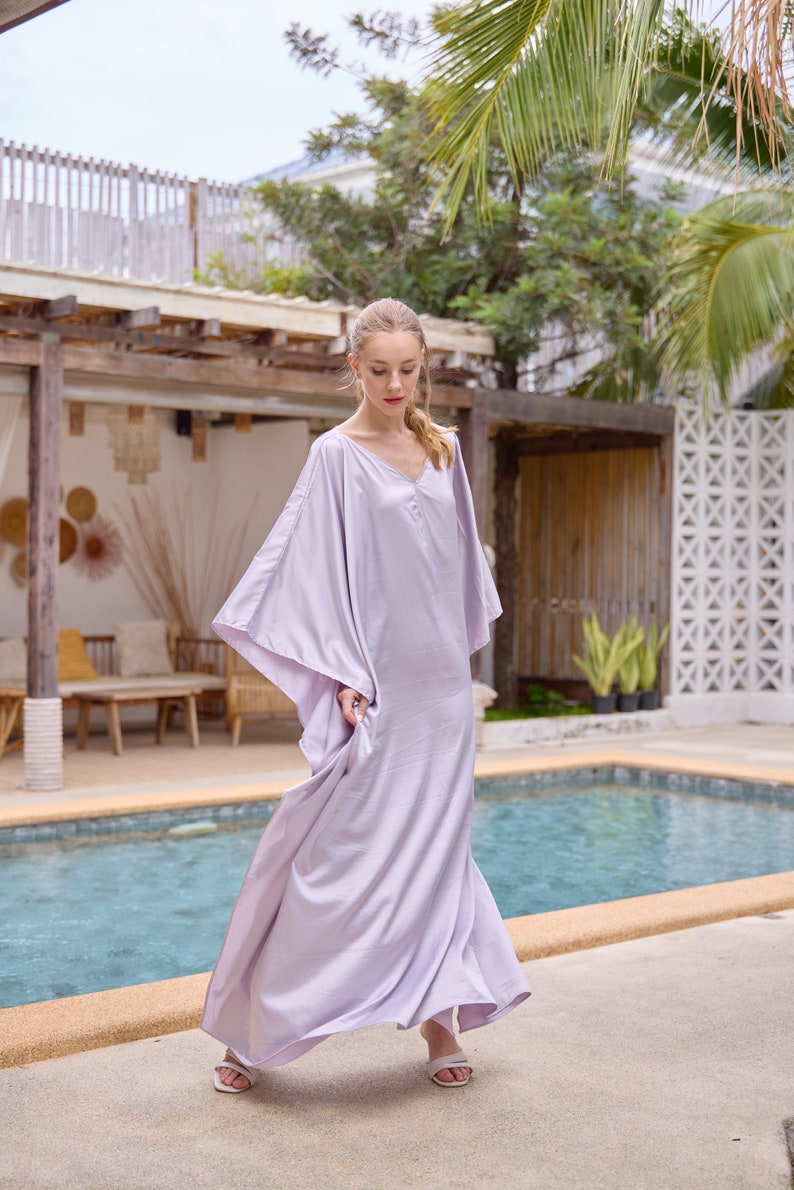 Puede incluir: Una mujer con un vestido maxi de color lavanda fluido con mangas largas y cuello en V. Est&aacute; de pie junto a una piscina.