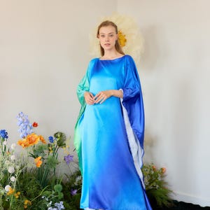 May include: A woman wearing a long, flowing blue and green ombre dress with a wide neckline. The dress has a loose, flowing fit and is made of a silky fabric. The woman is standing in front of a white wall with flowers in the foreground.