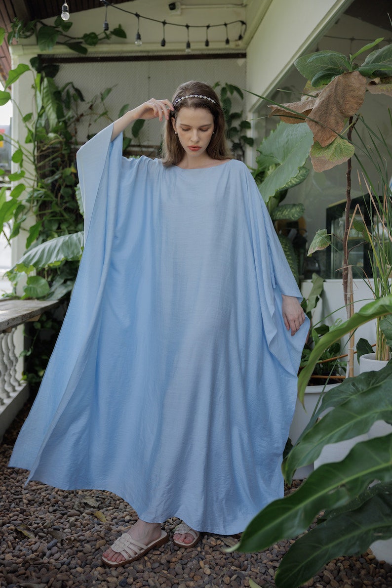 K&ouml;nnte beinhalten: Ein hellblaues Maxikleid mit einer lockeren, flie&szlig;enden Silhouette. Das Kleid hat einen runden Ausschnitt und lange &Auml;rmel. Das Model tr&auml;gt das Kleid mit einem Stirnband und Sandalen.