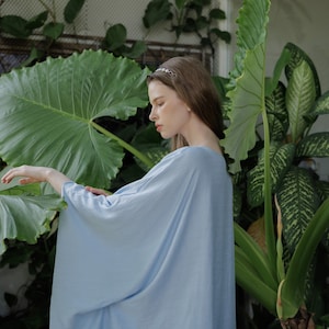 K&ouml;nnte beinhalten: Eine Frau in einem hellblauen, flie&szlig;enden, lang&auml;rmeligen Kleid steht in einem &uuml;ppigen gr&uuml;nen Garten. Sie tr&auml;gt ein silbernes Stirnband und hat lange braune Haare.
