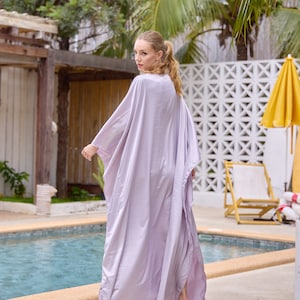 Puede incluir: Una mujer con una larga t&uacute;nica de color lila claro y fluida se encuentra junto a una piscina. La t&uacute;nica tiene un escote amplio y abierto y mangas largas. La mujer est&aacute; mirando por encima del hombro a la c&aacute;mara.