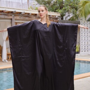 Peut inclure: Un caftan noir fluide avec un col en V. Le v&ecirc;tement a des manches larges et une coupe ample. Le mod&egrave;le porte des sandales blanches. L'arri&egrave;re-plan comprend une piscine et un feuillage tropical.