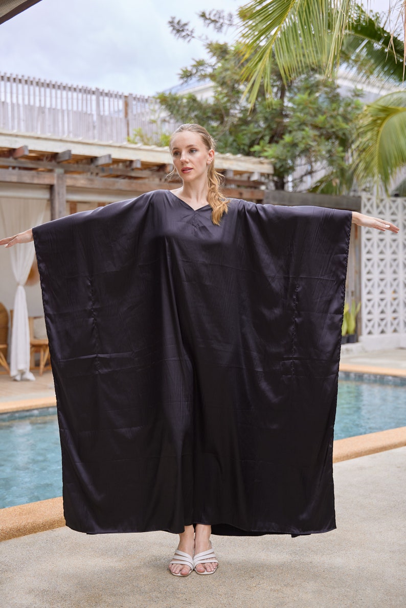 Peut inclure: Un caftan noir fluide avec un col en V. Le v&ecirc;tement a des manches larges et une coupe ample. Le mod&egrave;le porte des sandales blanches. L'arri&egrave;re-plan comprend une piscine et un feuillage tropical.