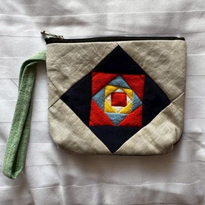 Puede incluir: Una pequeña bolsa de tela beige con cierre de cremallera y una correa verde. La bolsa presenta un diseño de patchwork colorido con un cuadrado central rojo, azul, amarillo y blanco.