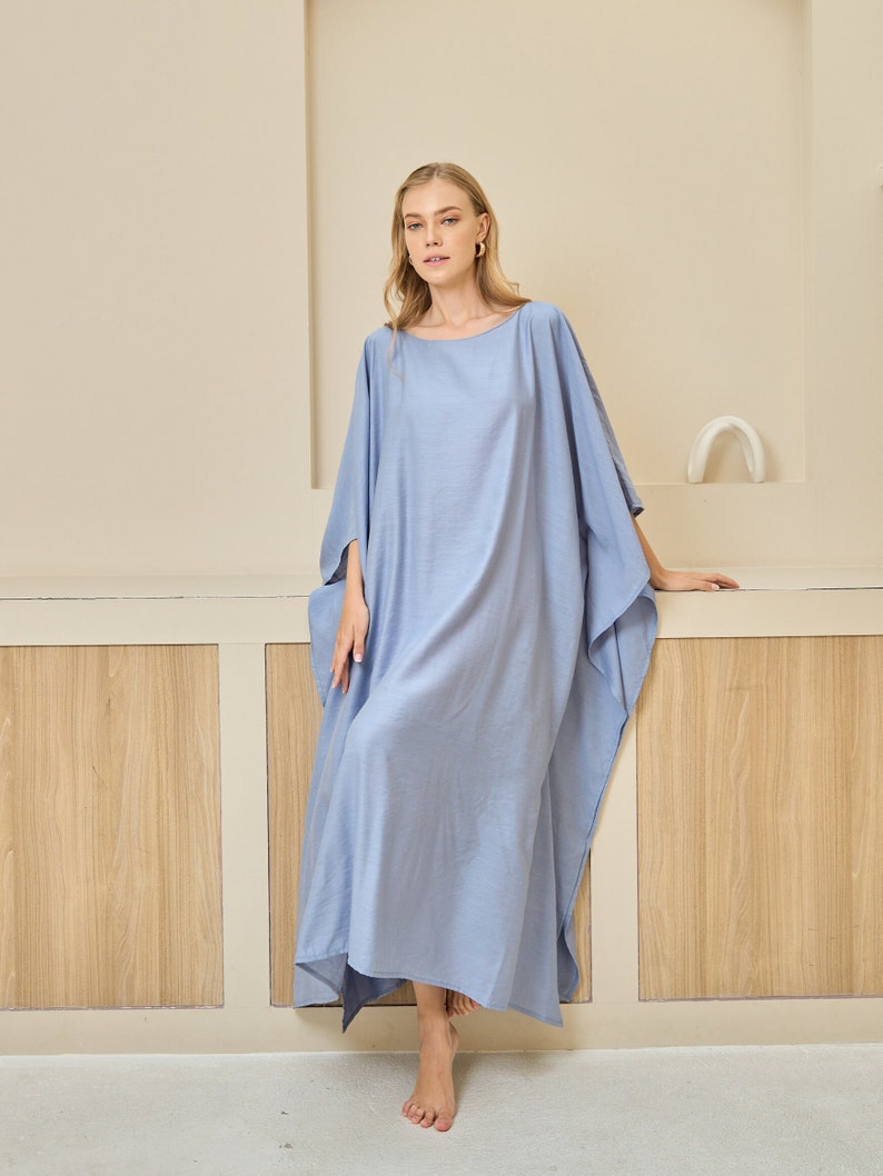 May include: A floor-length, light blue kaftan dress featuring a wide, loose-fitting silhouette with batwing sleeves and a simple round neckline. The garment is made of a solid-colored, lightweight fabric that drapes down to the ankles.