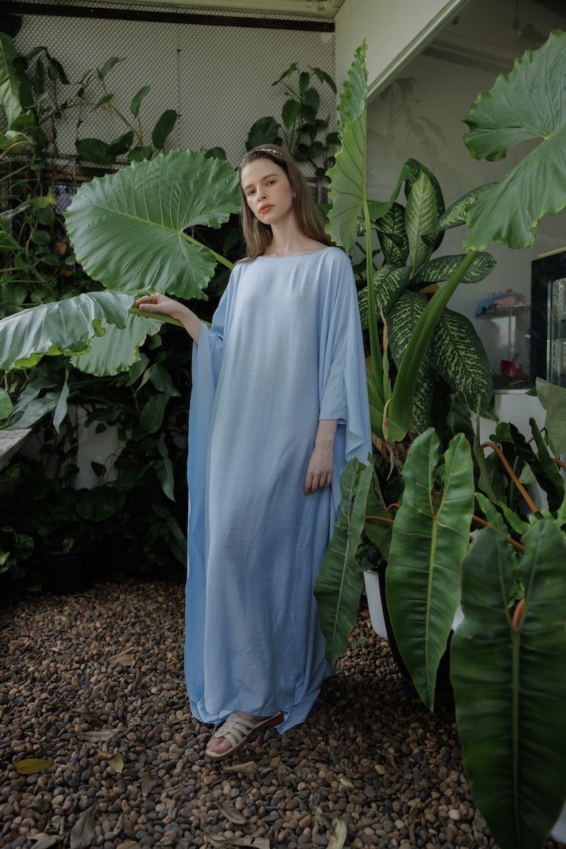 K&ouml;nnte beinhalten: Eine Frau tr&auml;gt ein hellblaues Maxikleid mit flie&szlig;ender Silhouette. Sie steht in einem Garten mit &uuml;ppigem Gr&uuml;n.