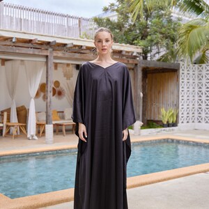 Peut inclure: Une femme porte une robe kaftan noire, ample, avec un col en V. La robe est longue et a des manches larges. Elle porte des sandales blanches. L'arri&egrave;re-plan comprend une piscine et de la v&eacute;g&eacute;tation tropicale.