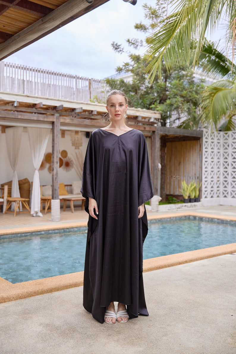 Peut inclure: Une femme porte une robe kaftan noire, ample, avec un col en V. La robe est longue et a des manches larges. Elle porte des sandales blanches. L'arri&egrave;re-plan comprend une piscine et de la v&eacute;g&eacute;tation tropicale.