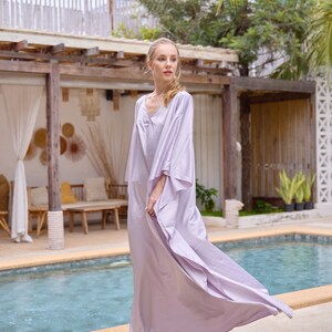 Puede incluir: Una mujer con un vestido maxi de color lavanda fluido con cuello en V y mangas anchas, de pie junto a una piscina.