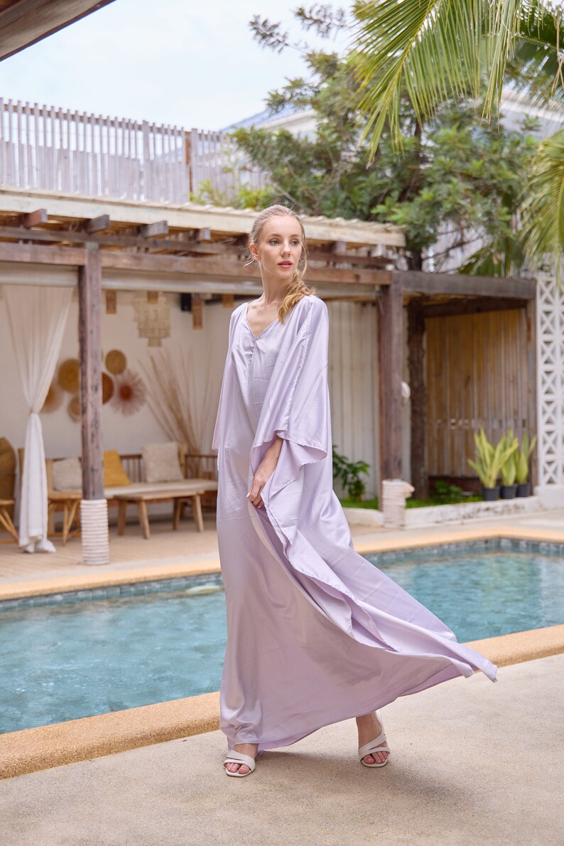 Puede incluir: Una mujer con un vestido maxi de color lavanda fluido con cuello en V y mangas anchas, de pie junto a una piscina.