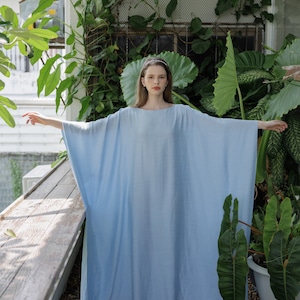 K&ouml;nnte beinhalten: Eine Frau tr&auml;gt ein hellblaues, locker sitzendes Maxikleid mit langen &Auml;rmeln. Sie steht in einer Gartenumgebung mit &uuml;ppigem Gr&uuml;n.