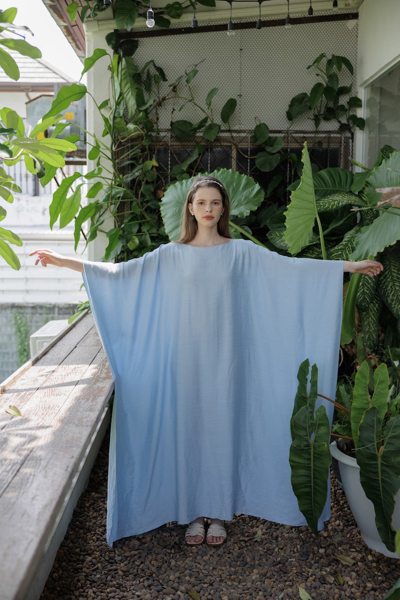 K&ouml;nnte beinhalten: Eine Frau tr&auml;gt ein hellblaues, locker sitzendes Maxikleid mit langen &Auml;rmeln. Sie steht in einer Gartenumgebung mit &uuml;ppigem Gr&uuml;n.