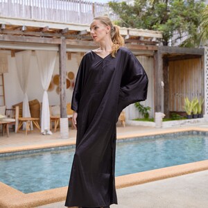 Peut inclure: Une longue robe caftan noire avec un col en V et des manches larges. La robe est faite d'une mati&egrave;re brillante et soyeuse et arrive au sol. Le mod&egrave;le porte des sandales blanches. L'arri&egrave;re-plan comprend une piscine et des plantes tropicales.