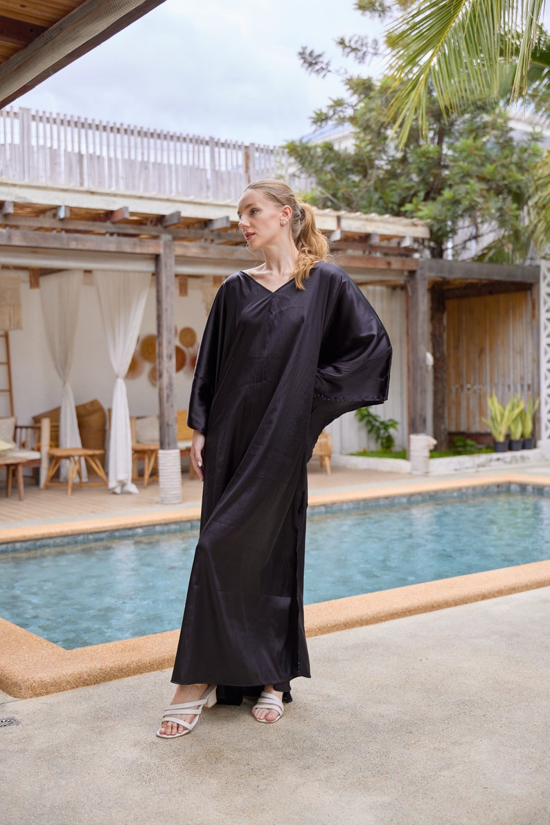 Peut inclure: Une longue robe caftan noire avec un col en V et des manches larges. La robe est faite d'une mati&egrave;re brillante et soyeuse et arrive au sol. Le mod&egrave;le porte des sandales blanches. L'arri&egrave;re-plan comprend une piscine et des plantes tropicales.