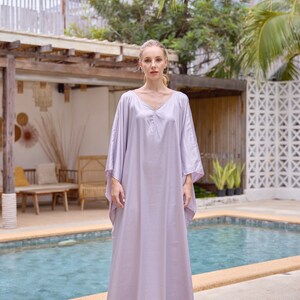 Puede incluir: Una mujer con un vestido largo de caft&aacute;n de color lila claro y fluido. Est&aacute; de pie junto a una piscina con una palmera en el fondo.