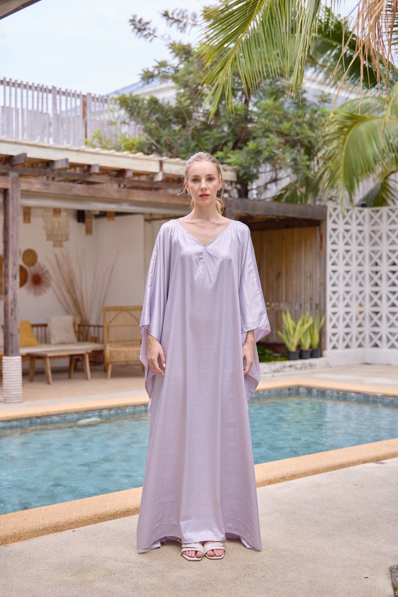 Puede incluir: Una mujer con un vestido largo de caft&aacute;n de color lila claro y fluido. Est&aacute; de pie junto a una piscina con una palmera en el fondo.