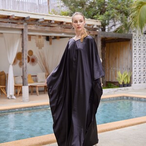 Peut inclure: Longue robe noire fluide avec un col en V. La robe a des manches larges et une coupe ample. La personne porte des sandales blanches. L'arri&egrave;re-plan comprend une piscine et un feuillage tropical.