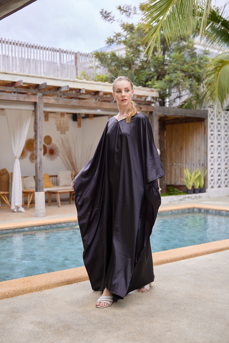 Peut inclure: Longue robe noire fluide avec un col en V. La robe a des manches larges et une coupe ample. La personne porte des sandales blanches. L'arri&egrave;re-plan comprend une piscine et un feuillage tropical.