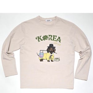Puede incluir: Una camiseta beige de manga larga con un estampado gráfico de un dachshund con un sombrero y ropa tradicional coreana. El texto "KOREA" está impreso en verde sobre el perro.