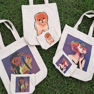 May include: Four tote bags with different designs. The first bag has a cartoon dog with a pink nose and tongue. The second bag has a cartoon woman with rainbow hair blowing a bubble. The third bag has a cartoon woman with brown hair and a yellow shirt. The fourth bag has a cartoon woman with brown hair and a yellow shirt.