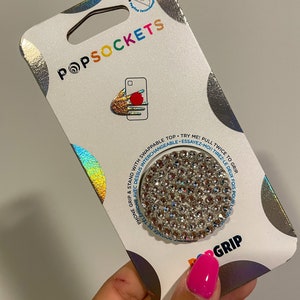 May include: A PopSockets phone grip and stand with a circular, silver, rhinestone-covered top. The packaging is white with the PopSockets logo and text in multiple colors. The grip is designed to be swappable.