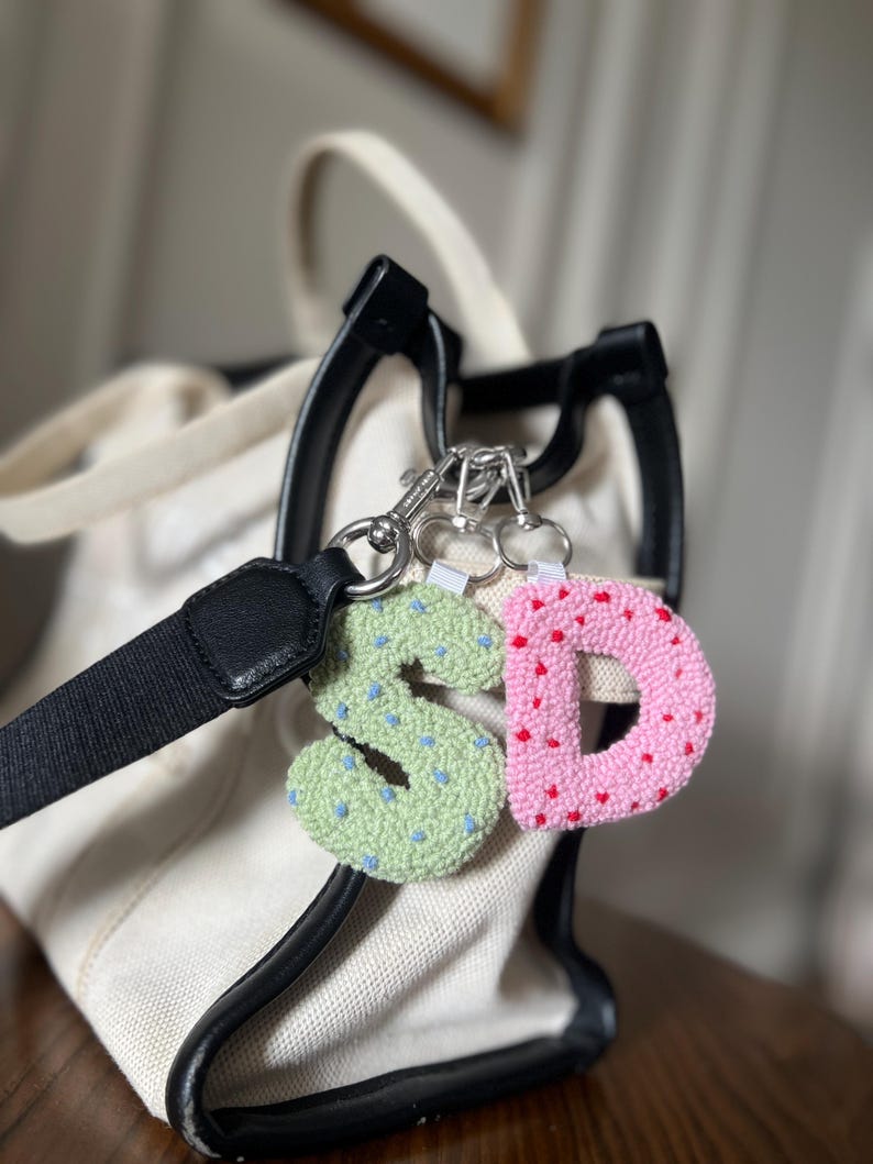 May include: Two letter keychains, featuring the letters "S" and "D". The "S" is light green with blue dots, and the "D" is pink with red dots. The keychains are attached to a silver clasp, with a black and white bag in the background.