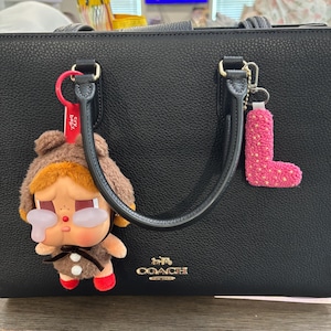 May include: Black Coach handbag with gold hardware and a top handle. A brown plush doll with red shoes and a pink letter "L" keychain hang from the handle. The Coach logo is visible on the front.