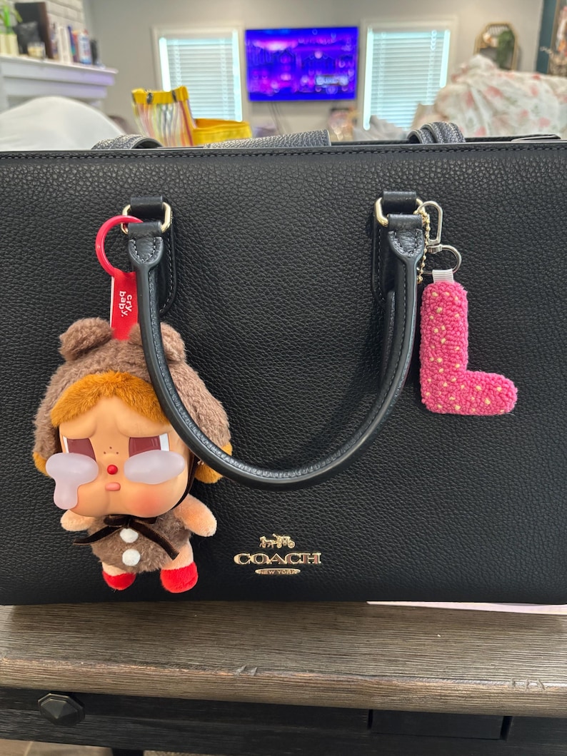 May include: Black Coach handbag with gold hardware and a top handle. A brown plush doll with red shoes and a pink letter "L" keychain hang from the handle. The Coach logo is visible on the front.