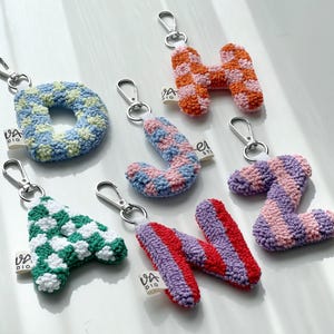 May include: A collection of handmade letter keychains in various colors and patterns. The letters include D, H, A, J, N, and Z, each with a silver-tone clasp. The keychains are made of a textured, possibly crocheted, material.