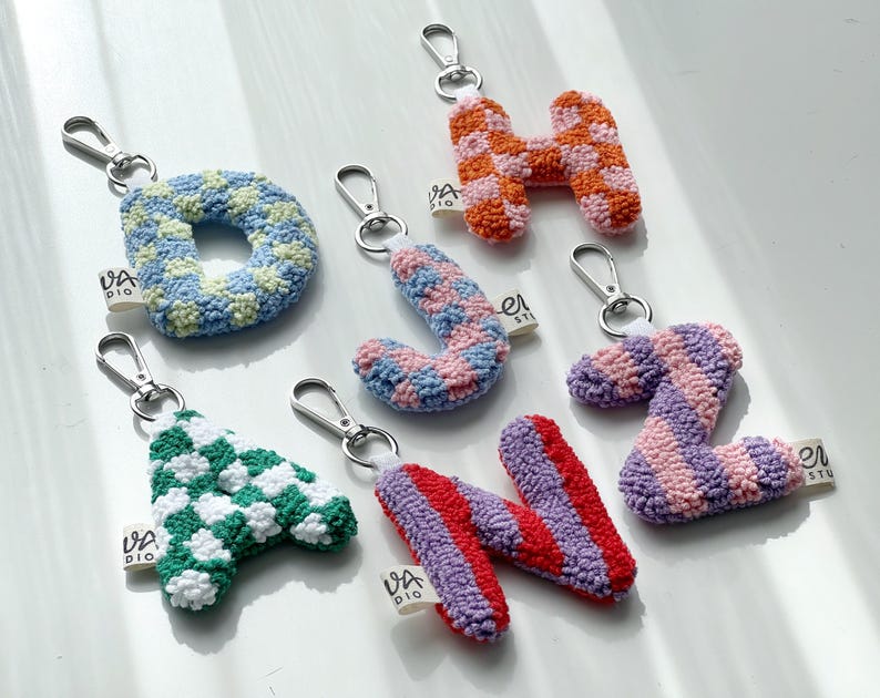 May include: A collection of handmade letter keychains in various colors and patterns. The letters include D, H, A, J, N, and Z, each with a silver-tone clasp. The keychains are made of a textured, possibly crocheted, material.
