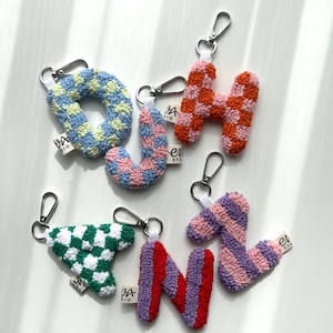 May include: A collection of handmade letter keychains in various colors and patterns. The letters include O, H, J, N, and a triangle shape, each with a silver-tone clasp. The keychains feature checkered and striped designs in shades of blue, pink, orange, green, red, and purple.