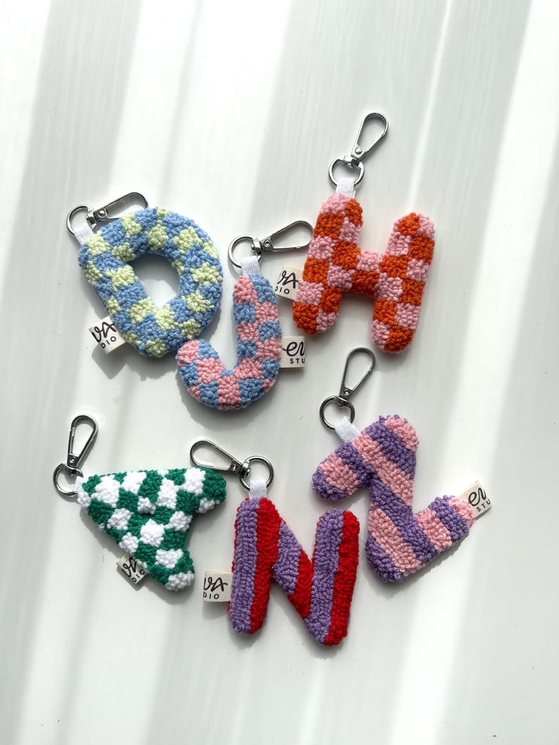 May include: A collection of handmade letter keychains in various colors and patterns. The letters include O, H, J, N, and a triangle shape, each with a silver-tone clasp. The keychains feature checkered and striped designs in shades of blue, pink, orange, green, red, and purple.