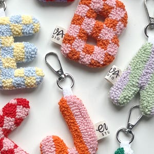May include: A collection of colorful, handmade letter keychains. The letters are made of a textured, woven material in various patterns and colors, including orange and pink checkerboard, and striped orange and pink. Each keychain has a silver clasp.