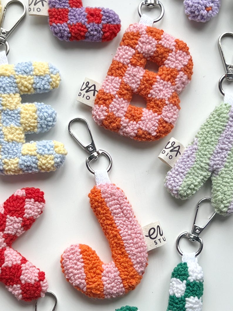 May include: A collection of colorful, handmade letter keychains. The letters are made of a textured, woven material in various patterns and colors, including orange and pink checkerboard, and striped orange and pink. Each keychain has a silver clasp.