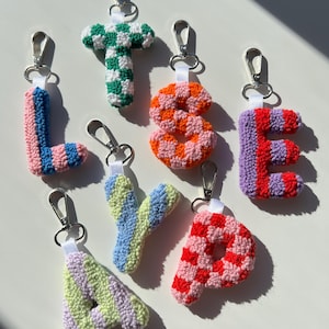 May include: A collection of handmade, colorful crochet letter keychains. Each letter is a different color combination, including pink and blue, green and white, orange and pink, red and purple, and red and pink. They have silver-tone clasps.