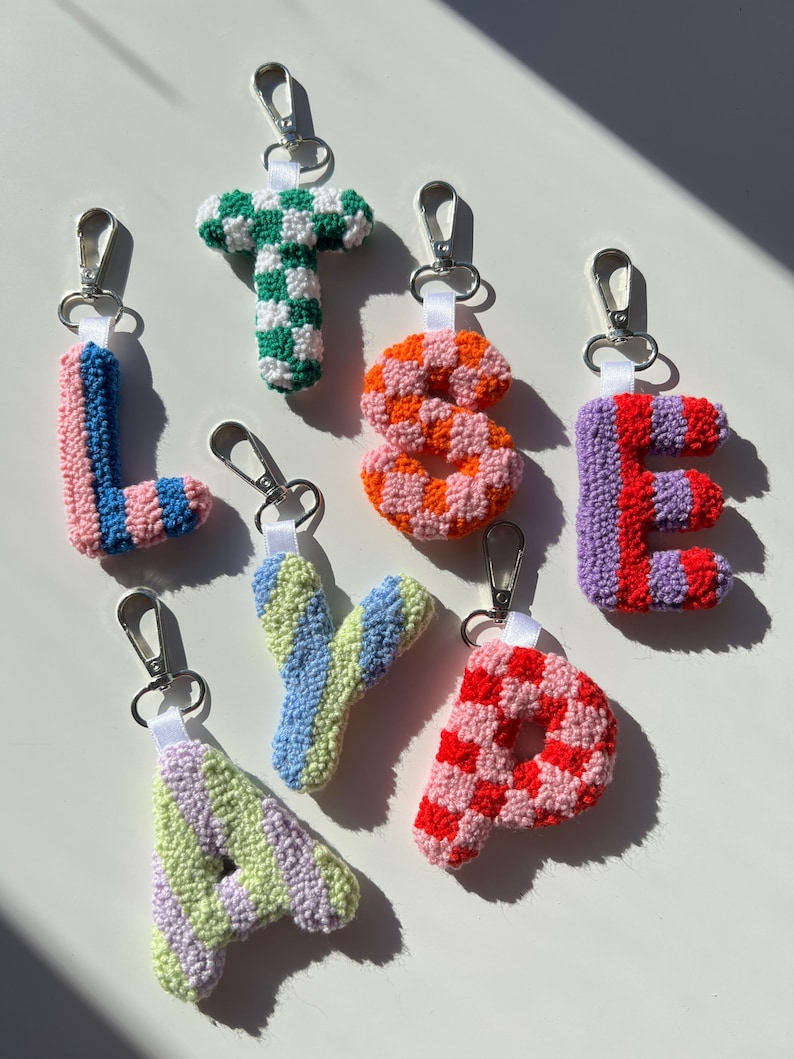 May include: A collection of handmade, colorful crochet letter keychains. Each letter is a different color combination, including pink and blue, green and white, orange and pink, red and purple, and red and pink. They have silver-tone clasps.