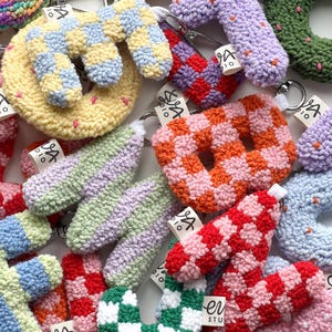 May include: A collection of colorful, handmade crochet keychains in various shapes, including letters and donuts. The keychains feature a variety of patterns, such as checkerboard and polka dots, and are attached to silver key rings. Each keychain has a small tag.