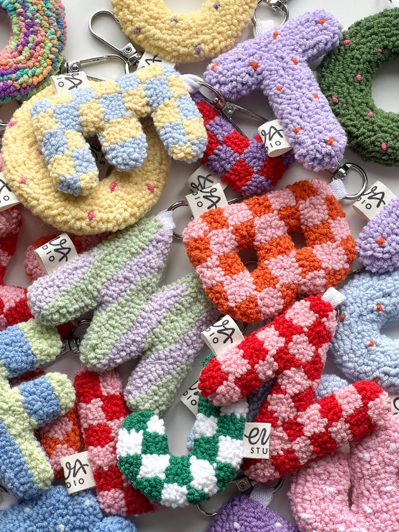 May include: A collection of colorful, handmade crochet keychains in various shapes, including letters and donuts. The keychains feature a variety of patterns, such as checkerboard and polka dots, and are attached to silver key rings. Each keychain has a small tag.